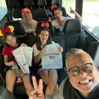 Group of smiling people in a van, some wearing Mickey Mouse ears, holding souvenir bags.