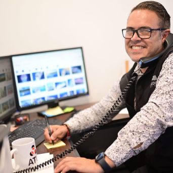 Person smiling at desk, talking on phone, typing on computer, and taking notes.