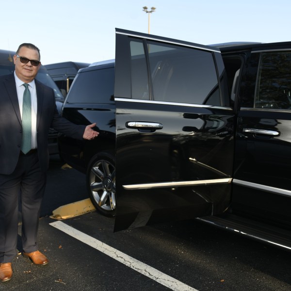 Man in suit stands next to open door of black SUV in parking lot.