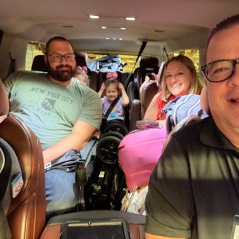 Four people smiling inside a car, with luggage visible.