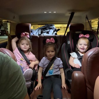 Three young children with bows in their hair sit in car seats inside a vehicle.